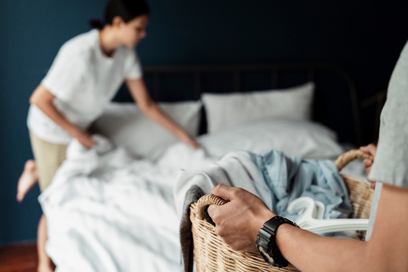 Someone brings in a basket of laundry while a woman makes the bed