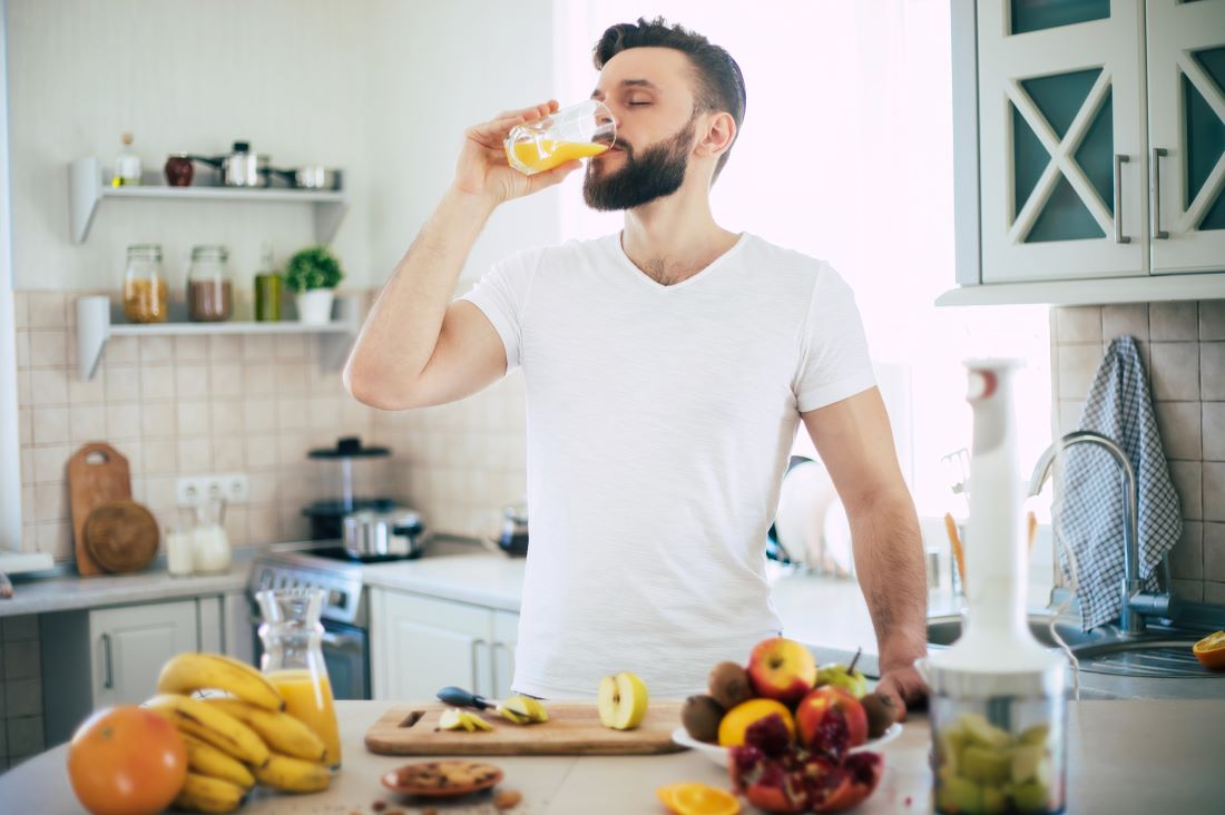 Man drinking orange juice with a bunch of fruits on the counter.